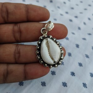 May include: A silver pendant featuring a white cowrie shell. The shell is set in a decorative silver frame with a beaded border. The pendant has a loop for a chain.