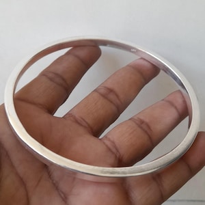Pode incluir: Uma pulseira de bracelete prateada brilhante. A pulseira redonda é segurada em uma mão e o metal reflete a luz. A pulseira tem um design simples e elegante, adequado para uso diário ou ocasiões especiais.
