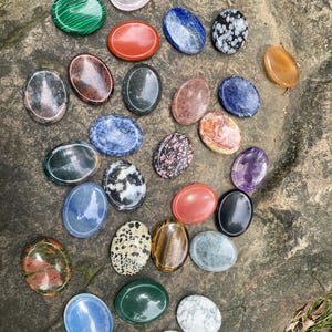 Könnte beinhalten: Eine Sammlung ovaler Edelsteine in verschiedenen Farben und Mustern, darunter grüner Malachit, roter Jaspis, blauer Lapislazuli und Tigerauge. Die Steine sind auf einer rauen, grauen Steinoberfläche angeordnet.