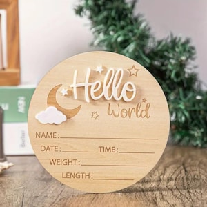 May include: A round wooden baby announcement sign with the words "Hello World" in white script. The sign includes spaces to write the baby's name, date, time, weight, and length. Decorative elements include a moon, cloud, and stars.