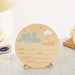 May include: A round wooden sign with the words "Hello World" in blue and brown. The sign has space to write a baby's name, date, time, weight, and length. It is decorated with a cloud, stars, and a moon.