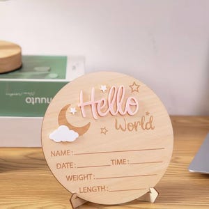 May include: A wooden, round baby announcement sign with the words "Hello World" in pink. The sign features a moon, cloud, and stars. It has lines for name, date, time, weight, and length.