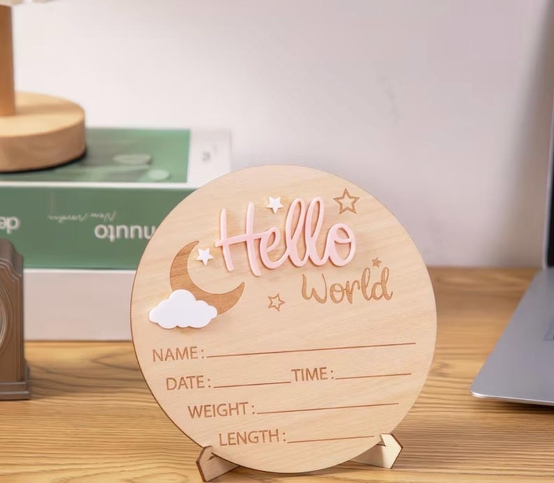 May include: A wooden, round baby announcement sign with the words "Hello World" in pink. The sign features a moon, cloud, and stars. It has lines for name, date, time, weight, and length.