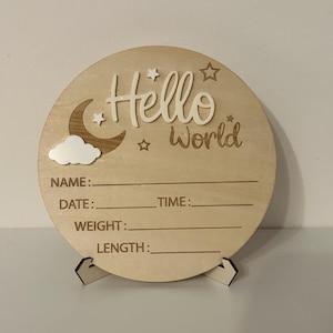 May include: A round wooden sign with the words "Hello World" in a script font, along with a moon, stars, and cloud design. The sign has spaces to record a baby's name, date, time, weight, and length.