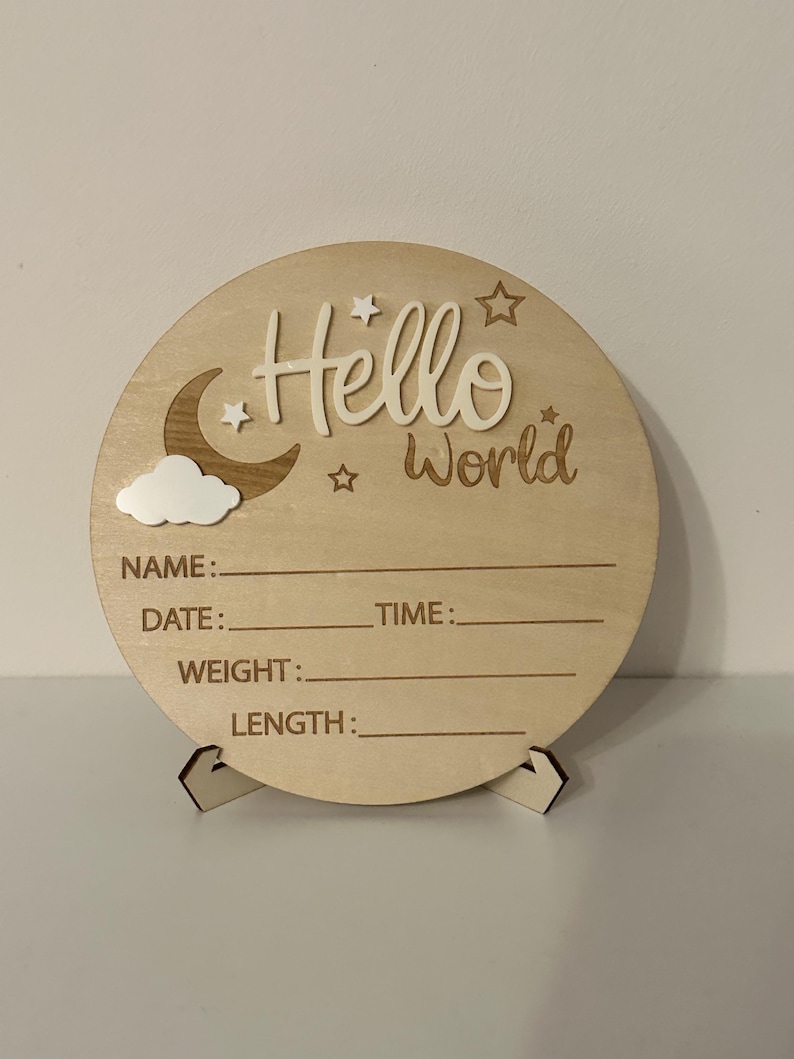 May include: A round wooden sign with the words "Hello World" in a script font, along with a moon, stars, and cloud design. The sign has spaces to record a baby's name, date, time, weight, and length.