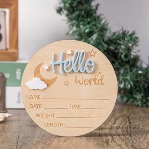 May include: A round wooden baby announcement sign with the words "Hello World" in blue and light brown. The sign includes spaces to write the baby's name, date, time, weight, and length. A crescent moon and cloud are also featured.