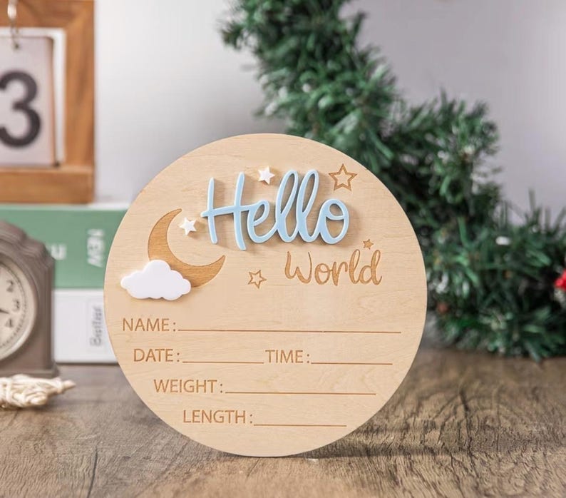 May include: A round wooden baby announcement sign with the words "Hello World" in blue and light brown. The sign includes spaces to write the baby's name, date, time, weight, and length. A crescent moon and cloud are also featured.