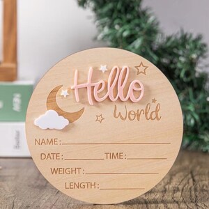 May include: A round wooden baby announcement sign with the words "Hello World" in pink and engraved text. The sign includes a moon, cloud, and star accents. It has spaces to write the baby's name, date, time, weight, and length.