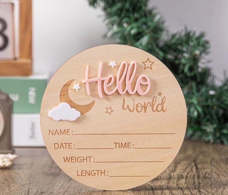 May include: A round wooden baby announcement sign with the words "Hello World" in pink and engraved text. The sign includes a moon, cloud, and star accents. It has spaces to write the baby's name, date, time, weight, and length.