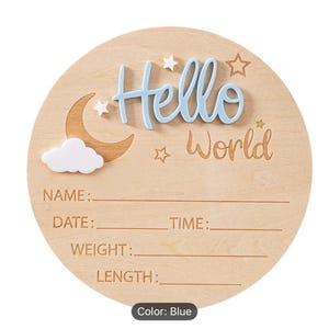 May include: A round wooden sign with the words "Hello World" in blue and brown. The sign has a moon, cloud, and star design. It includes spaces to write the baby's name, date, time, weight, and length.