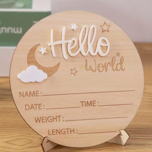 May include: A round wooden sign with the words "Hello World" in white script. The sign features a crescent moon, stars, and a cloud. It has lines for Name, Date, Time, Weight, and Length. The sign is displayed on a wooden stand.