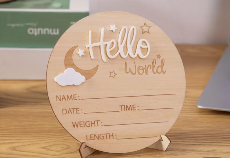 May include: A round wooden sign with the words "Hello World" in white script. The sign features a crescent moon, stars, and a cloud. It has lines for Name, Date, Time, Weight, and Length. The sign is displayed on a wooden stand.