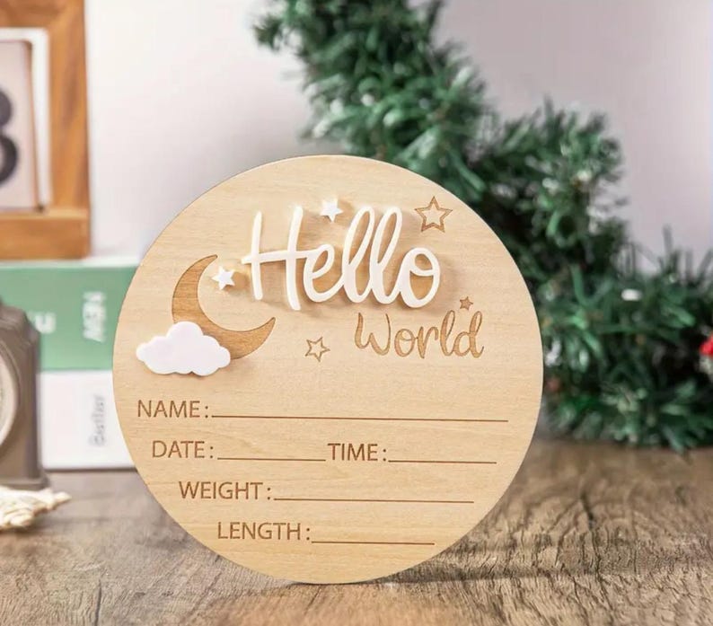 May include: A round wooden baby announcement sign with the words "Hello World" in white script. The sign includes spaces to write the baby's name, date, time, weight, and length. Decorative elements include a moon, cloud, and stars.