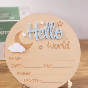 May include: A round wooden sign with the words "Hello World" in blue and brown. The sign has a moon, cloud, and star design. It includes lines for name, date, time, weight, and length. The sign is supported by a wooden stand.