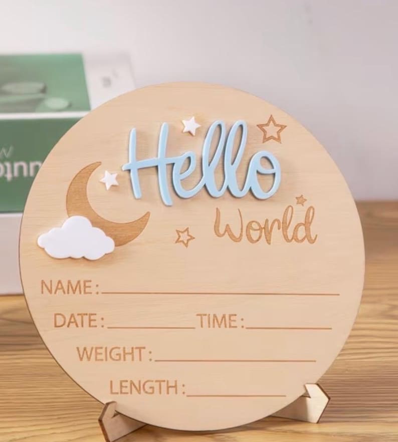 May include: A round wooden sign with the words "Hello World" in blue and brown. The sign has a moon, cloud, and star design. It includes lines for name, date, time, weight, and length. The sign is supported by a wooden stand.