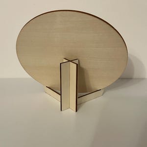 May include: A wooden, round sign with a light brown finish, supported by a matching wooden stand. The stand has a triangular base and a central support structure. The sign is displayed against a white background.