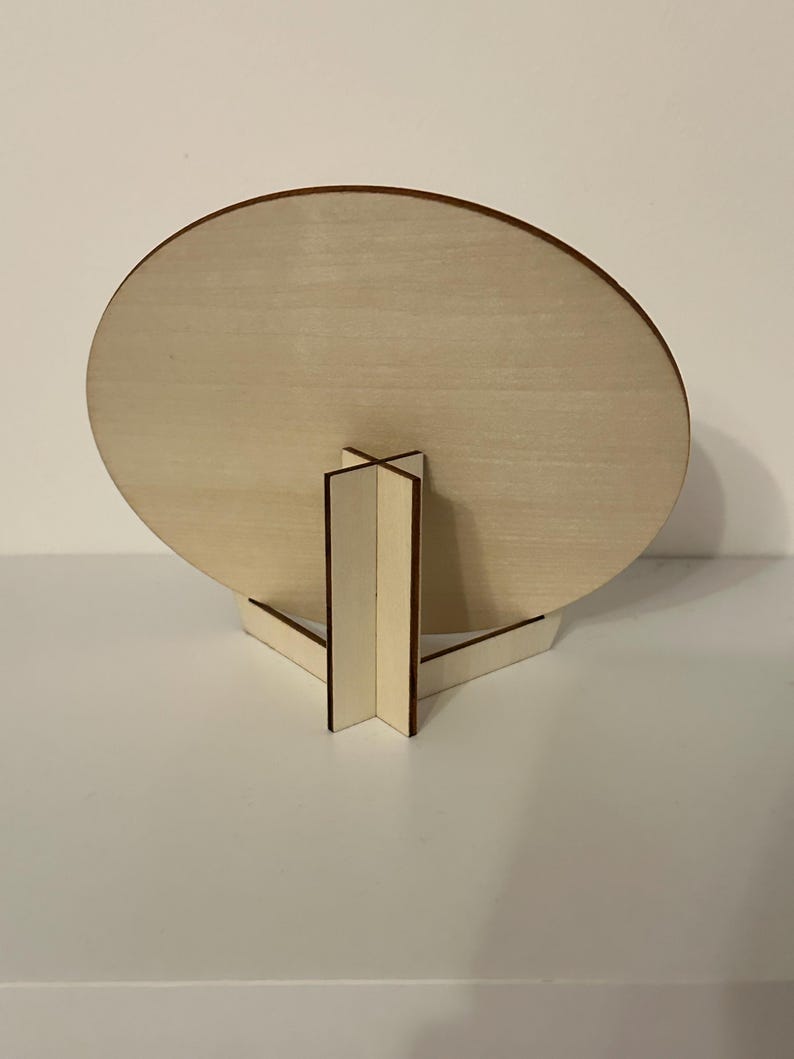 May include: A wooden, round sign with a light brown finish, supported by a matching wooden stand. The stand has a triangular base and a central support structure. The sign is displayed against a white background.