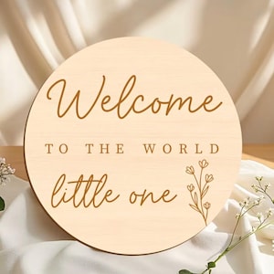 May include: A round wooden sign with the words "Welcome to the World little one" in brown script. A floral design is on the right side. The sign is light beige and sits on a white cloth.