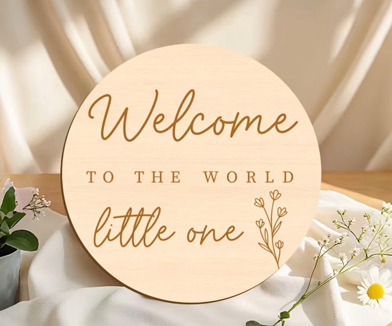 May include: A round wooden sign with the words "Welcome to the World little one" in brown script. A floral design is on the right side. The sign is light beige and sits on a white cloth.