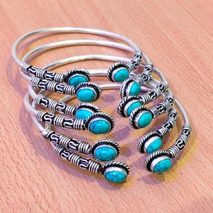 Peut inclure: Plusieurs bracelets en métal argenté ornés de pierres ovales turquoise. Les bracelets présentent un design complexe et sont disposés sur une surface en bois brun clair.