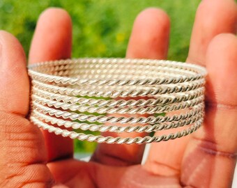 May include: A set of six silver bangles with a twisted rope pattern. The bangles are held in a hand, with a blurred green background. The bangles are made of a shiny metal.