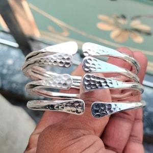May include: A collection of silver-coloured, hammered-texture wrap bracelets. Each bracelet has a spiral design with overlapping ends. The metal has a shiny finish, reflecting light. The bracelets are stacked together.