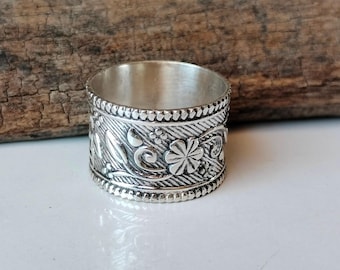 Könnte beinhalten: Ein silberner Ring mit einem aufwendigen Blumen- und Blattmuster. Der Ring hat ein breites Band, das mit detaillierten Schnitzereien von Blumen, Blättern und Wirbelmustern verziert ist. Die Oberfläche des Rings hat ein strukturiertes Aussehen mit poliertem Finish.