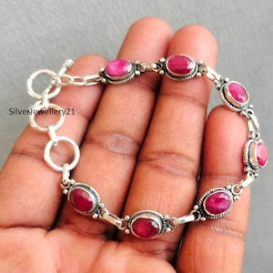 May include: A silver bracelet featuring oval-shaped, vibrant red gemstones. The bracelet is composed of interconnected links with each gemstone set in a decorative bezel. The clasp is a simple toggle style. The bracelet is displayed against a neutral background.