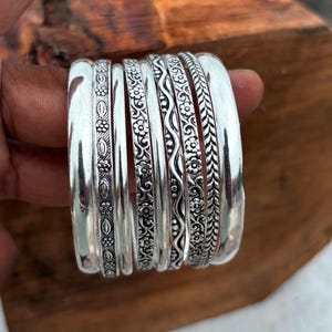May include: A set of seven silver-toned bangles featuring varied textures and patterns. The collection includes smooth polished bands, a band with a repeating floral motif, a wavy line design with dots, and a band with a braided or leaf-like pattern.