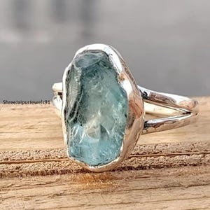 Könnte beinhalten: Ein Silberring mit einem großen, unregelmäßig geformten, hellblauen Edelstein. Der Ring hat ein geteiltes Schaftdesign und ein strukturiertes Band. Der Edelstein ist in einer polierten Silberfassung gefasst. Der Ring wird auf einer Holzoberfläche präsentiert.