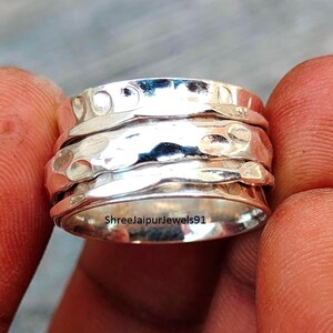 Peut inclure: Une bague argentée avec un motif texturé. La bague comporte plusieurs bandes, dont certaines avec une texture martelée. La bague est tenue entre deux doigts. Le texte "Shreejaipurjewels91" est visible sur la bague.