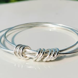 May include: Three silver bangles, joined by a decorative knot made of silver wire. The jewellery is presented on a white surface, reflecting light. The design is simple and elegant. The bangles are approximately 6 cm in diameter.