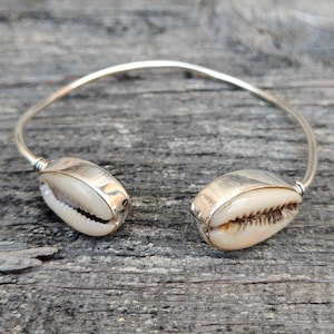 Puede incluir: Una pulsera de puño plateada con dos conchas de cauri naturales. Las conchas son de color crema con un interior marrón y están montadas en los extremos de la pulsera curva y abierta. La pulsera se exhibe sobre una superficie de madera envejecida.