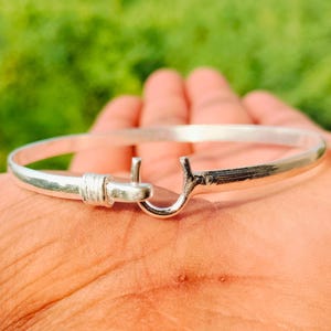Könnte beinhalten: Ein silberfarbenes Armreif-Armband mit Hakenverschluss. Das Armband ist rund und scheint aus Metall zu sein. Der Hakenverschluss befindet sich auf der rechten Seite des Armbands. Das Armband wird auf der Hand einer Person präsentiert.
