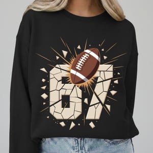 May include: Black sweatshirt featuring a brown and white football graphic bursting through the number "67". The number is designed to look like it is breaking apart. The football has white laces and is surrounded by a burst of yellow and orange.