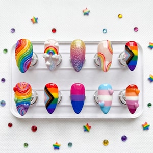 Pode incluir: Uma coleção de dez unhas artificiais com vários designs, incluindo arco-íris, glitter e padrões de bandeiras do orgulho. As unhas são exibidas em um suporte de plástico transparente, rodeadas por estrelas e gemas coloridas.