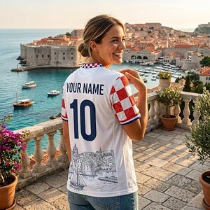 May include: A white short-sleeved jersey featuring a red and white checkered pattern on the sleeves, the text "YOUR NAME" in dark blue, the number "10" in large dark blue digits, and a black and white line drawing of a coastal city scene.