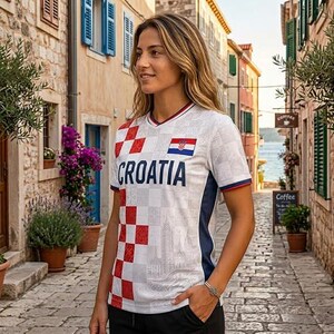 May include: A white V-neck soccer jersey featuring a red and white checkered pattern on the left side, the word CROATIA in dark blue letters, a small Croatian flag emblem on the chest, and dark blue side panels with a subtle architectural print.