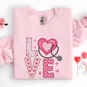 May include: A pale pink sweatshirt featuring the word "LOVE" in a decorative font. The "L" is patterned with swirls and hearts, the "O" is a heart with a stethoscope, and the "V" and "E" are covered in pink hearts. Pink stars are scattered around the word.