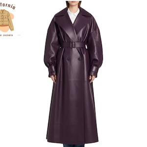 Puede incluir: Un abrigo largo de cuero morado oscuro con diseño cruzado y cinturón a juego. El abrigo presenta un cuello ancho y mangas largas. La modelo lleva una camisa blanca debajo. El abrigo es de California Leather Jackets.