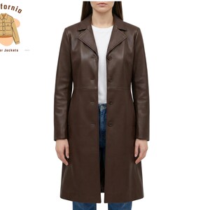 May include: A long, brown leather coat with a button-up front. The coat has a classic lapel collar and a tailored fit. The model is wearing a white t-shirt and blue jeans underneath. The coat is from California Leather Jackets.