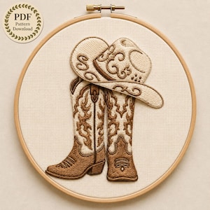 May include: An embroidery hoop containing a cream-colored fabric with a stitched design of a pair of brown and cream cowboy boots and a matching cowboy hat. A circular badge in the upper left corner displays the text PDF Pattern Download.