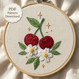 Puede incluir: Un bastidor de bordado que contiene una pieza de tela con un diseño bordado a mano de dos cerezas rojas, hojas verdes y dos flores blancas con centros amarillos, acentuado por pequeñas estrellas amarillas. El texto PDF Pattern Download es visible.