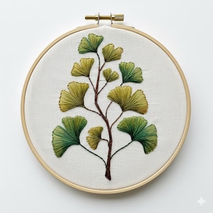 May include: An embroidered ginkgo leaf design on white fabric, set in a wooden embroidery hoop. The leaves are shades of green and yellow, with a brown stem. The hoop is secured with a brass screw.