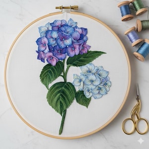 Op de afbeelding: Geborduurde hortensia bloem in een houten ring. De bloem heeft blauwe en paarse bloemblaadjes, groene bladeren en een stengel. Borduurgaren en een gouden schaar zijn zichtbaar.