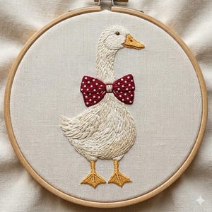 May include: Embroidered artwork of a goose wearing a red bow tie with white polka dots. The goose is white with yellow feet and beak, stitched on a cream-colored fabric, and framed in a wooden hoop. A charming, handmade piece.