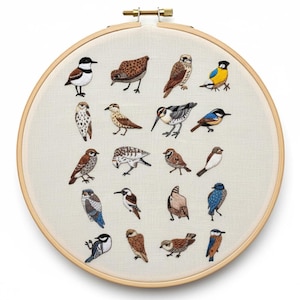Puede incluir: Un bastidor de bordado redondo con una colección de pájaros bordados en varios colores. Las aves están representadas con un estilo detallado y realista, con una variedad de poses y patrones de plumaje. El fondo es una tela beige claro, y el bastidor es de madera.