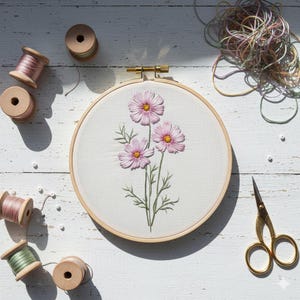 Può includere: Un telaio da ricamo con un design floreale con tre fiori di cosmo rosa con centri gialli e steli verdi. Il telaio è circondato da rocchetti di filo di legno in vari colori, insieme a un paio di forbici dorate.
