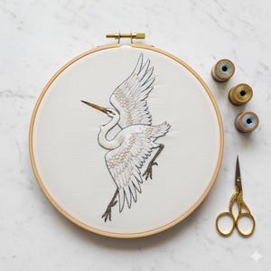 Peut inclure: Broderie d'un héron blanc en vol, sur tissu crème, dans un cercle à broder en bois. Les ailes de l'oiseau sont déployées, avec des détails de plumes. Ciseaux dorés et bobines de fil à côté du cercle.
