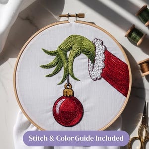 Pode incluir: Um bastidor de bordado circular de madeira contendo tecido branco com um desenho bordado &agrave; m&atilde;o de uma m&atilde;o verde de dedos longos com uma manga vermelha de borda branca, segurando um enfeite de Natal vermelho. Texto: Stitch & Color Guide Included.